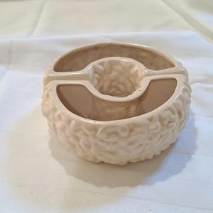 May include: A beige ceramic ashtray with a textured, rounded exterior. The ashtray has a central circular holder and two side rests. The design is simple and functional, suitable for home or office use.