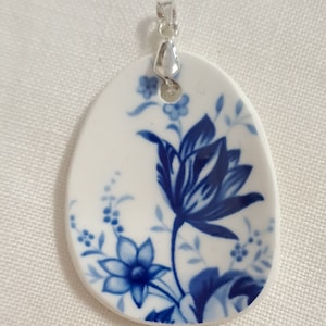 May include: White oval pendant with a blue floral design. The pendant features a large blue flower and smaller floral accents. A silver bail is attached at the top. The pendant is likely ceramic or porcelain.
