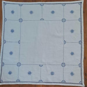 May include: A square white fabric cloth featuring a dark blue embroidered grid pattern. The design includes small, intricate star-like floral motifs centred within each grid square and a decorative border along the outer edges of the fabric.