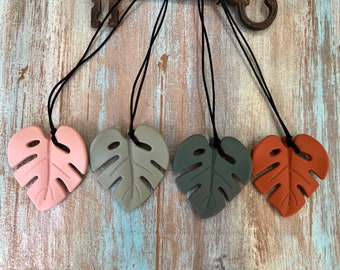 May include: Four silicone monstera leaf necklaces in pink, gray, dark gray, and orange. Each necklace has a black cord.