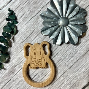May include: A wooden teether shaped like an elephant with a smiling face and a fish in its mouth. The teether is light brown and has a smooth, rounded surface.