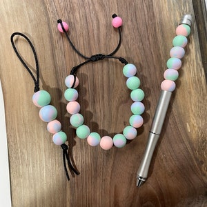 May include: A set of three handmade accessories featuring pastel pink, blue, and green marbled beads. The set includes a bracelet, a keychain, and a pen.