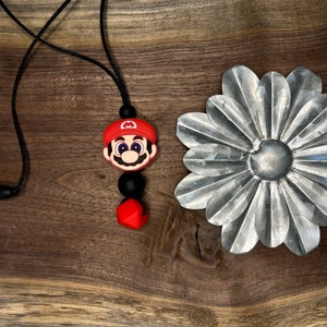 May include: A black and red silicone necklace with a Mario character charm. The necklace is on a wooden surface.