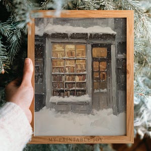 Könnte beinhalten: Ein gerahmter Druck einer winterlichen Buchladen-Szene. Das Kunstwerk zeigt ein schneebedecktes Gebäude mit einem warm beleuchteten Fenster, das Bücherregale zeigt. Der Rahmen ist aus hellem Holz gefertigt, und der Text "MY PRINTABLE JOY" ist sichtbar.