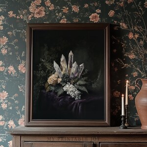 Puede incluir: Una impresión enmarcada que presenta una naturaleza muerta de cristales, helechos y flores blancas sobre una tela morada oscura. La obra de arte está ambientada sobre un papel pintado floral oscuro. Un mueble de madera y velas están en primer plano.