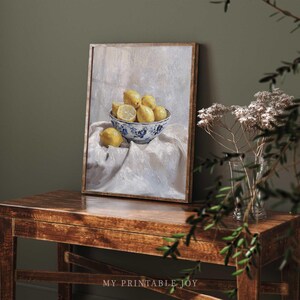 May include: A painting of a bowl of lemons on a white tablecloth. The bowl is blue and white with a floral design. The lemons are yellow and some are cut in half. The painting is framed in a light brown frame.