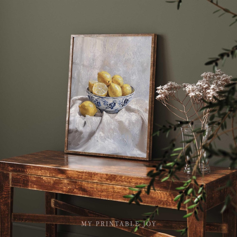 May include: A painting of a bowl of lemons on a white tablecloth. The bowl is blue and white with a floral design. The lemons are yellow and some are cut in half. The painting is framed in a light brown frame.