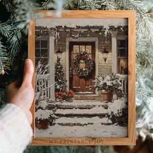 Puede incluir: Una impresión enmarcada de una escena navideña nevada. La obra de arte representa un porche con una corona en la puerta, un pequeño árbol de Navidad y plantas cubiertas de nieve. El marco es de madera clara. El texto "MY PRINTABLE JOY" es visible.