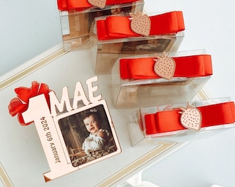 May include: A rose gold photo frame with the name "MAE" and the number "1" with the date "January 6th 2024". Clear boxes with red ribbon and strawberry accents are also visible.