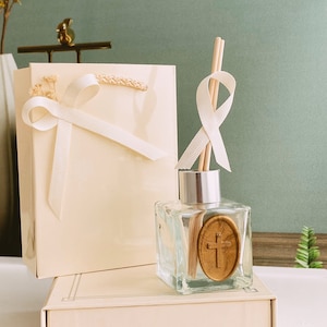 Puede incluir: Una bolsa de regalo blanca con una cinta blanca y una botella de vidrio transparente con una cruz de madera en la parte delantera. La botella está sentada sobre una caja blanca con una tapa blanca. El fondo es una pared azul claro.