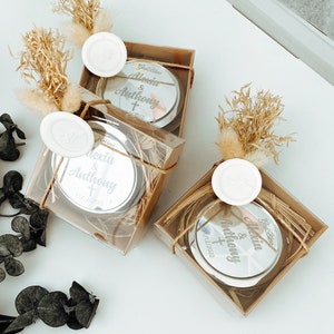 Könnte beinhalten: Drei kleine, transparente Geschenkboxen mit braunen Papier- und Bindfadenakzenten. Jede Box enthält einen runden, silbernen Kompaktspiegel mit einer personalisierten Inschrift, die "God Bless Alexia & Anthony" und ein Datum lautet. Die Boxen sind mit getrockneten Blumen und einem weißen Wachssiegel verziert.