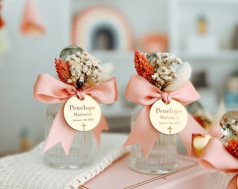 May include: Clear glass bottles with a textured design, decorated with pink ribbons, dried flowers, and gold tags. The tags read "Penelope Mattesich January 6th 2024" with a cross symbol. The bottles are arranged on a pink box.