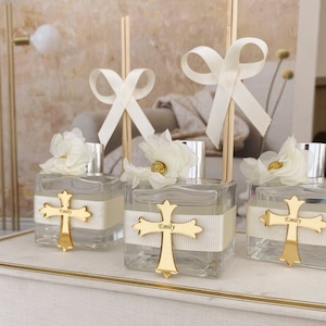 Może przedstawiać: Trzy przezroczyste butelki szklane z złotymi zawieszkami w kształcie krzyża i białymi akcentami wstążki. Każda butelka ma biały kwiat i drewniany patyczek z białą kokardką. Butelki są ułożone na białym pudełku ze złotą obwódką. Butelki mają napis "Emily" na wstążce.