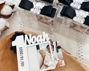 May include: White wooden frame with a photo of a baby boy and the text "Noah" and "09/16/2023". The frame is decorated with a white number one and a small white elephant figurine. The frame is sitting on a wooden surface.