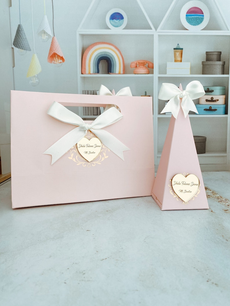 May include: Two pink gift boxes with white bows and gold heart-shaped tags that say "Isla Florence Dawn" and "Mr. Jackson".