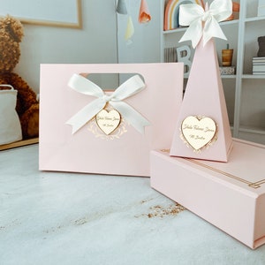 May include: Two pink gift boxes with gold accents. One box is a bag with a white bow and a heart-shaped tag that says "Isla Welcome Home". The other box is a pyramid shape with a white bow and a heart-shaped tag that says "Isla Welcome Home".