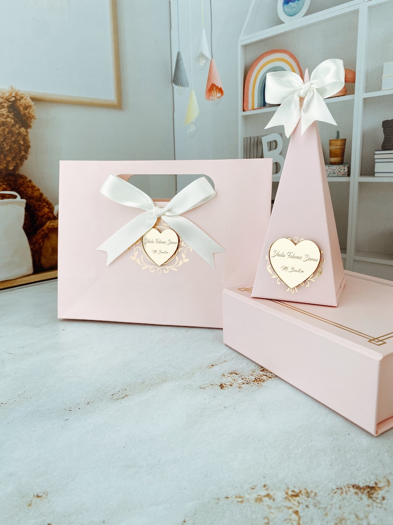 May include: Two pink gift boxes with gold accents. One box is a bag with a white bow and a heart-shaped tag that says "Isla Welcome Home". The other box is a pyramid shape with a white bow and a heart-shaped tag that says "Isla Welcome Home".