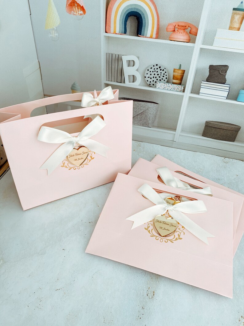 May include: Two pink gift bags with white satin ribbon bows and gold heart-shaped tags that say "Hola Bienvenida Dora" with a gold floral design.