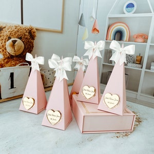 May include: Pink pyramid-shaped boxes with white bows and gold heart-shaped labels that say "Isla Welcome Home" in cursive. The boxes are arranged on a light grey surface.