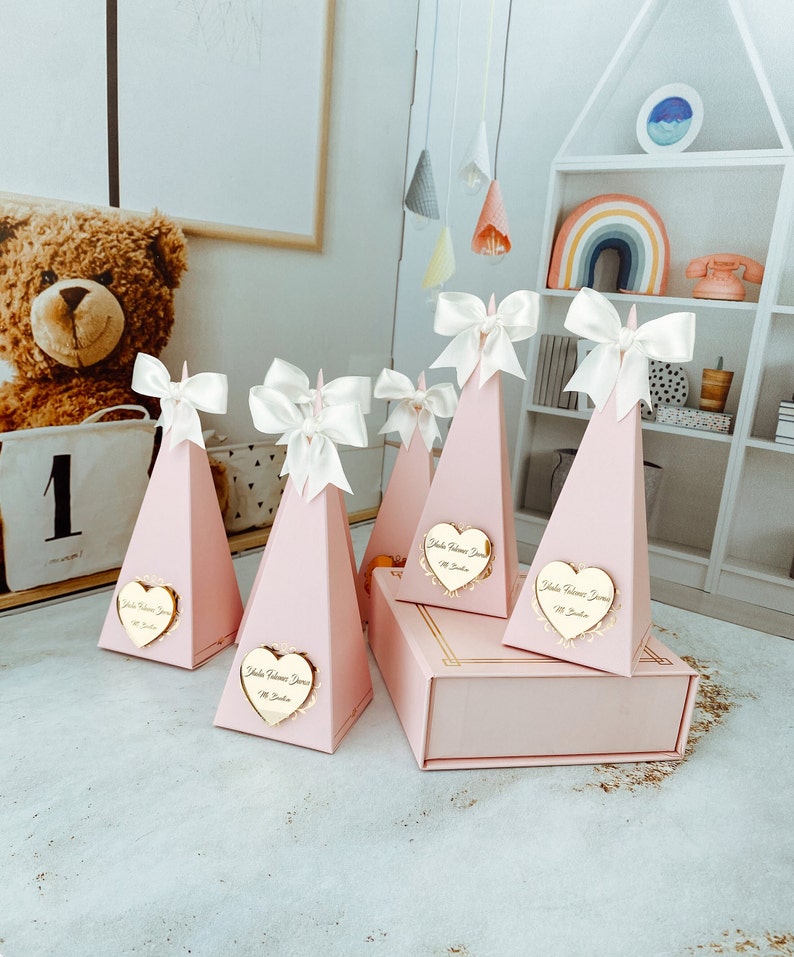 May include: Pink pyramid-shaped boxes with white bows and gold heart-shaped labels that say "Isla Welcome Home" in cursive. The boxes are arranged on a light grey surface.