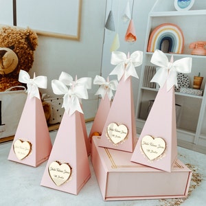 May include: Pink pyramid-shaped boxes with white satin bows and gold heart-shaped tags that say "Hola Bienvenida" with a small flower design. The boxes are arranged on a pink surface.
