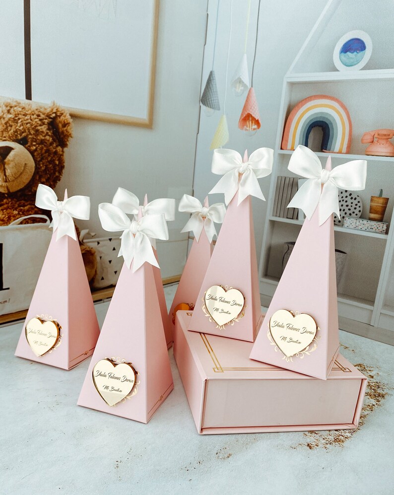 May include: Pink pyramid-shaped boxes with white satin bows and gold heart-shaped tags that say "Hola Bienvenida" with a small flower design. The boxes are arranged on a pink surface.