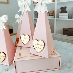 May include: Pink pyramid-shaped boxes with white bows and gold heart-shaped tags with the text "Isla Valencia Duran" and "11. Bautista". The boxes are arranged in a pyramid shape on a pink box with gold trim.