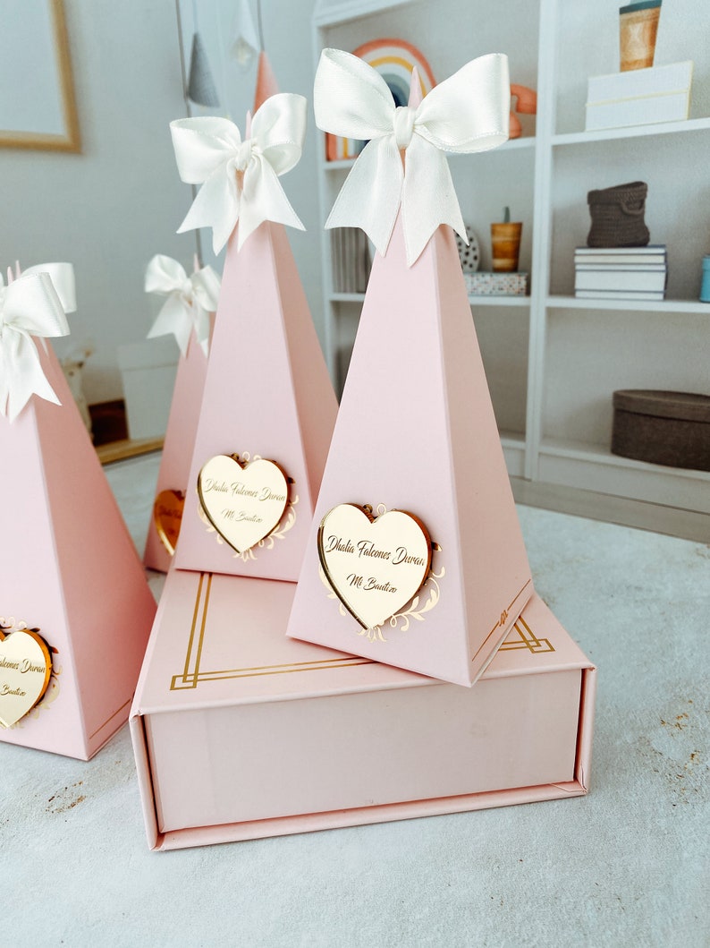 May include: Pink pyramid-shaped boxes with white bows and gold heart-shaped tags with the text "Isla Valencia Duran" and "11. Bautista". The boxes are arranged in a pyramid shape on a pink box with gold trim.