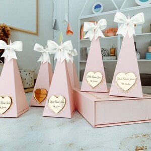 May include: Pink pyramid-shaped gift boxes with white bows and gold heart-shaped labels that say "Isla Welcome Home" in cursive. The boxes are arranged on a table with a white shelf unit in the background.