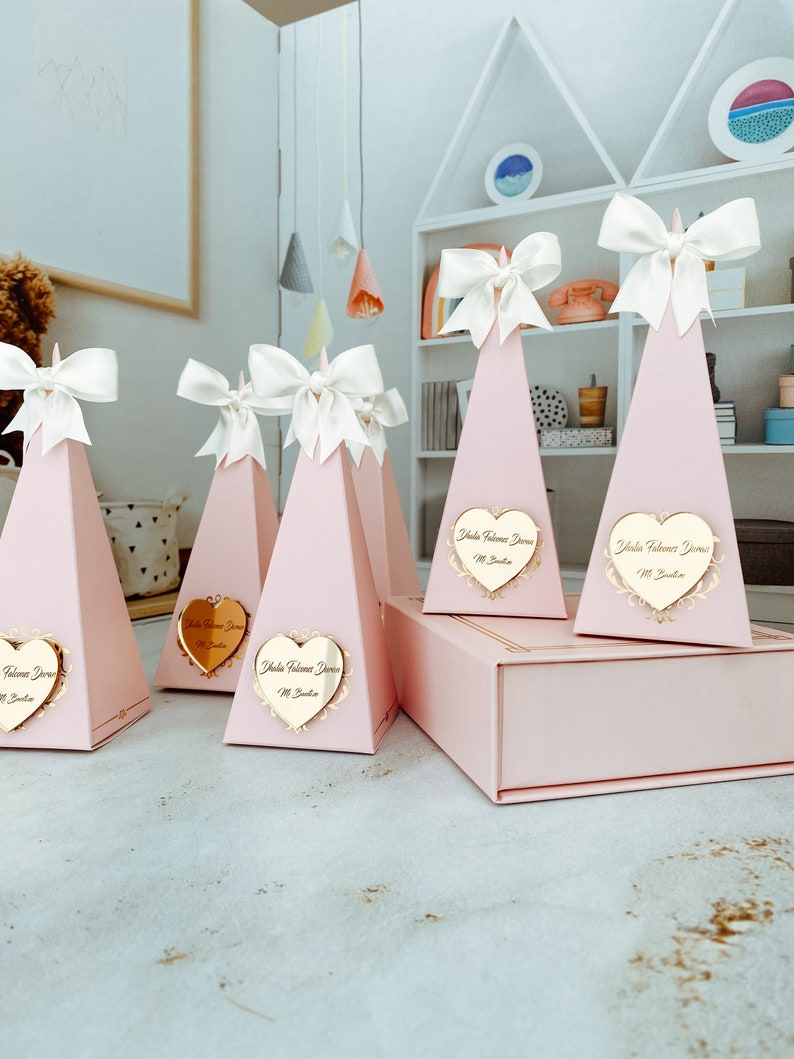 May include: Pink pyramid-shaped gift boxes with white bows and gold heart-shaped labels that say "Isla Welcome Home" in cursive. The boxes are arranged on a table with a white shelf unit in the background.