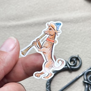 May include: A sticker featuring a medieval-style illustration of a brown dog playing a wind instrument. The dog wears a blue and white hat and a red collar. The sticker has a white border and is held by a person's fingers.