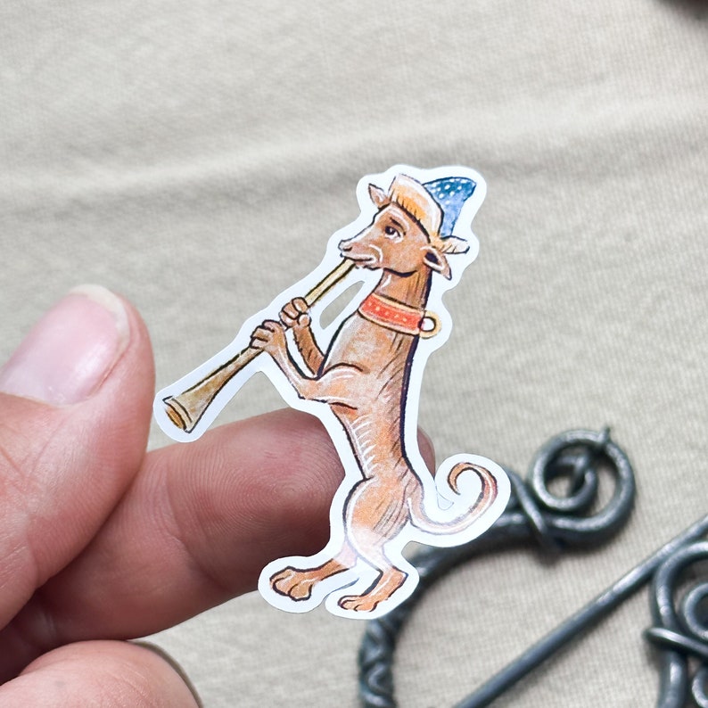 May include: A sticker featuring a medieval-style illustration of a brown dog playing a wind instrument. The dog wears a blue and white hat and a red collar. The sticker has a white border and is held by a person's fingers.