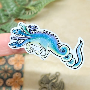 May include: A sticker featuring a multi-headed dragon in shades of blue and green. The dragon has a long, curved tail and a detailed, stylized design. The sticker is on a white background.