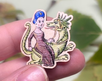 May include: A wooden pin with a hand-painted image of a woman in a blue hat riding a green dragon. The woman is wearing a purple dress and the dragon has a brown bridle.