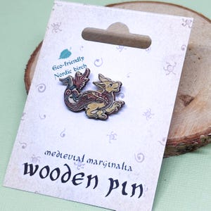 Op de afbeelding: Een houten pin met een middeleeuwse draak met rode, gele en groene details. De pin is gemaakt van milieuvriendelijk Scandinavisch berkenhout en is gelabeld "medieval marginalia wooden pin".