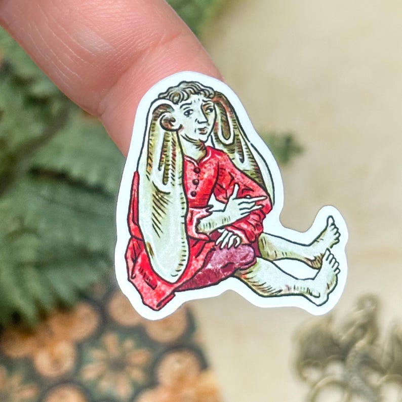 May include: A sticker featuring a medieval-style illustration of a figure with large, rabbit-like ears. The figure is dressed in a red tunic and seated with legs extended. The artwork has a vintage, hand-drawn aesthetic.