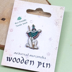 Puede incluir: Un pin de madera con una ilustración medieval de un hombre con una túnica y un sombrero verdes, sosteniendo un bastón. El pin está hecho de abedul nórdico ecológico y está etiquetado como "medieval marginalia wooden pin".
