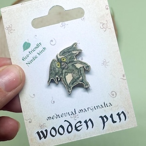 May include: A wooden pin featuring a green and white illustration of a winged creature with large eyes. The pin is on a card that says "medieval marginalia wooden pin" and "eco-friendly nordic birch".