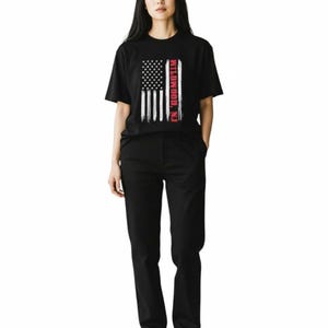 May include: A person wearing a black t-shirt with an American flag design and the text "Wildwood, NJ". The person is also wearing black pants and white sneakers. The outfit is casual and simple.