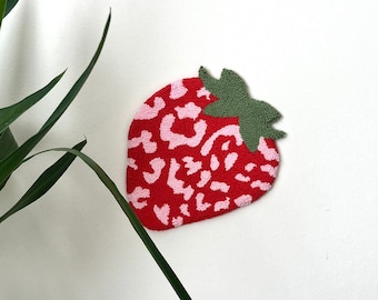 May include: A red and pink strawberry-shaped wall hanging with a leopard print design. The strawberry has green leaves.