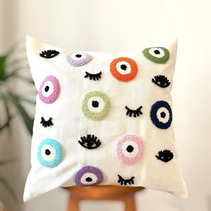 May include: White throw pillow with colorful embroidered eyes and eyelashes. The eyes are in various colors, including blue, green, pink, and orange.