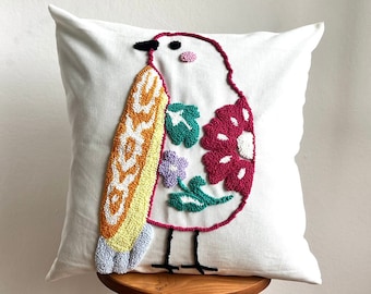 May include: White pillow with a colorful embroidered bird design. The bird has a yellow, orange, and white body, green leaves, and a pink flower.