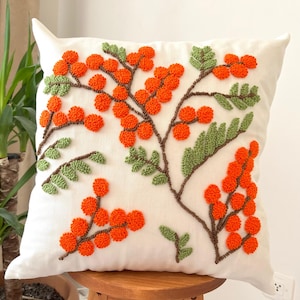 Puede incluir: Almohada blanca con un diseño floral naranja y verde. El diseño presenta una rama con bayas naranjas y hojas verdes. La almohada está hecha de una tela suave y mullida.