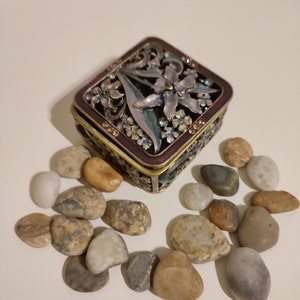 May include: A decorative, square jewelry box with a purple border and floral design. The box is surrounded by small, smooth stones in various shades of brown, gray, and white. The box has a hinged lid and intricate metalwork.