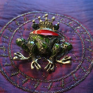 May include: A green and gold frog figurine with a red crown and green gem eyes. The frog is sitting on a purple fabric background with a decorative circle pattern.