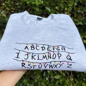 May include: A light gray crewneck sweatshirt featuring black embroidered letters of the alphabet arranged in three rows. Each row is connected by a black line with small, colorful embroidered light bulbs interspersed between the letters.
