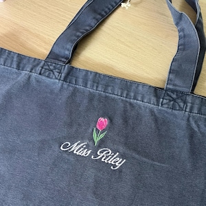 Könnte beinhalten: Eine anthrazitfarbene Canvas-Tragetasche mit einer Stickerei einer einzelnen rosa Tulpe mit grünen Blättern, die über dem weißen, in Schreibschrift gestickten Text Miss Riley platziert ist. Die Tasche hat zwei passende Stoffhenkel.
