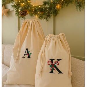 May include: Two cream-coloured drawstring gift bags, each embroidered with a letter. One features an "A" with green leaves and red berries, the other a "K" with candy canes and holly. A festive garland with lights and pine cones is visible in the background.
