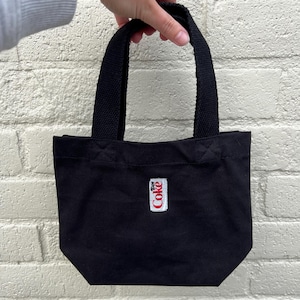 K&ouml;nnte beinhalten: Eine kleine schwarze Tragetasche aus Segeltuch mit zwei stabilen gewebten Griffen. Mittig auf der Vorderseite befindet sich ein rechteckiger wei&szlig;er Aufn&auml;her mit dem rot-schwarzen Diet Coke Logo. Die Tasche hat ein schlichtes, strukturiertes Design.