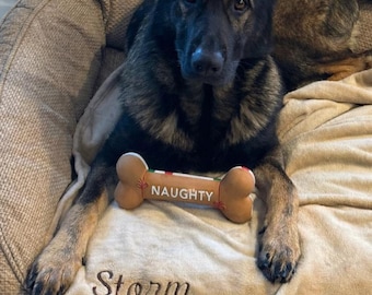 May include: A sable German Shepherd dog lying on a tan pet bed with a tan blanket. The dog is holding a tan, bone-shaped toy with the word NAUGHTY printed in white. The blanket is embroidered with the name Storm and a small paw print icon.